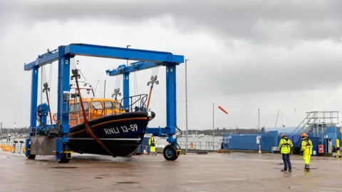 A lifeboat hangs on an orange strop from a blue frame. Workers in high visability jackets stand on the right of a brick paved pier. In the background murky skies hover over a marina with several boats and yachts. 