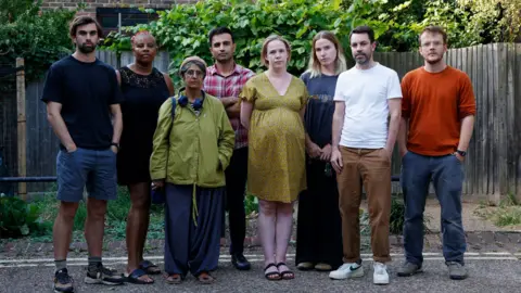 Eight residents from the Consort estate in Peckham stand in a line on a road near their properties. 