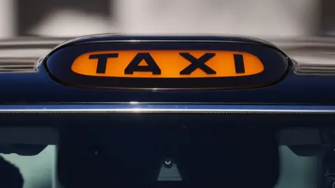 A general view of a taxi sign on top of a black cab. The taxi sign is orange with black font. 