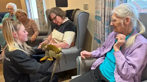 Kimberley Piper/BBC Ruth is sat on the right of the image and is one of four residents in the photo. They are all looking at a handler who is kneeling in front of them and holding a squirrel monkey.