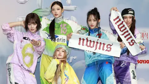 The five members of the K-pop music group NewJeans pose for the camera while wearing tracksuits and holding banners that say 'nwjns', pictured at the SBS Music Awards Summer at INSPIRE Arena in Jung-gu on July 21, 2024 in Incheon, South Korea.