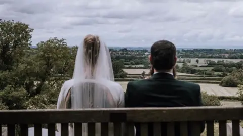 Oobaloos Photography A bride and groom sit side-by-side on a wooden bench with their backs to the camera, looking at the view of fields. The groom is wearing a dark suit. The bride is wearing a white dress. 