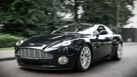 A black Aston Martin Vanquish on a road, with trees, blurred in the background
