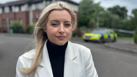 Sinn Fein MLA Órlaithí Flynn at the scene of a death in Dunmurry. She has long blonde hair and is wearing a black top and a white blazer.