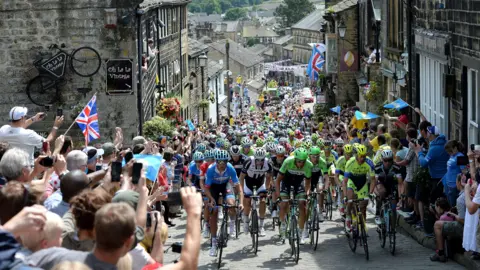 Cyclists climbing up a narrow street which has stone buildings on either side. Crowds line the street, a few holding Union Jack flags. There is bunting above the street further down the street which is the bottom of the road.