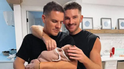 Family handout Adam Frisby and Jamie Corbett are cuddling their baby daughter Leven who is wrapped in a pink knitted blanket. 