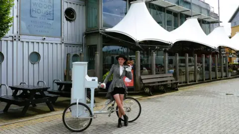 Chloe Marie Aston Chloe Marie Aston stands next to her piano bike outside a modern, steel-framed restaurant at a marina in Portishead, Bristol. She is wearing a black and white striped outfit with a black top hat and black boots.