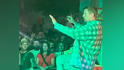 Cleah Hetherington/BBC A singer performs onstage. He has a shirt on and is holding a microphone in his right hand. He is standing in front of a crowd.