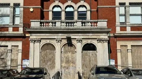 Alexander Barty A large, red brick building has its doors and windows boarded up. Cars are parked to the front of the building.