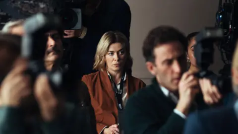 ITV Myles sat in the audience of a police conference room wearing a rusty orange blazer, looking intently past the camera. People with cameras are sat around her.