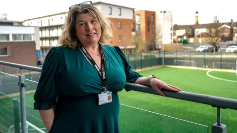 A woman with long blonde hair, glasses on top f her head, a green dress and a lanyard, smiles as she leans on a railing. She is standing on a balcony with a 3G football pitch behind her.