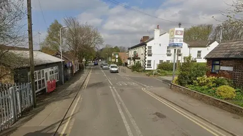 Google Station Road in Maghull on a dry day with Maghull Railway Station on the right and some cars travelling up the road.