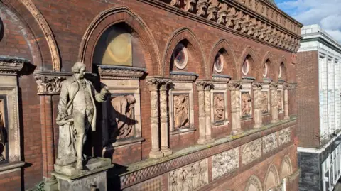 Stoke-on-Trent City Council An external side view of a building, with a statue of a man on a plinth, and detailed depictions of people in the walls behind