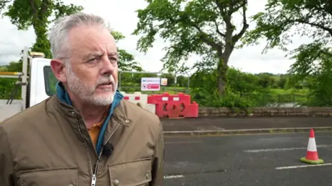 Guy Shackleton, with grey hair and beard, wears an olive green jacket and blue fleece and orange polo top as he stands in the street with the so-called royal lime trees and the River Ribble in the background.