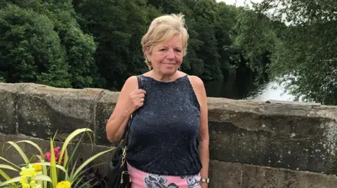 Pat Russell Russell stands on a bridge with a river behind her. She is in a sparkly black top and smiling.