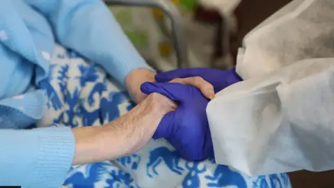 PA Media Two pairs of hands – one, wearing blue latex gloves, is the carer's, the others are bare and belong to the person being cared for. The hands are clasped together in a show of reassurance.