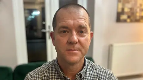 A head-and-shoulders shot of a man wearing a check shirt, looking expressionless directly at the camera.