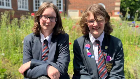 Jon Wright/BBC Two teenage school pupils wearing ties and blazers sit in front of a wild flowers and a brick building