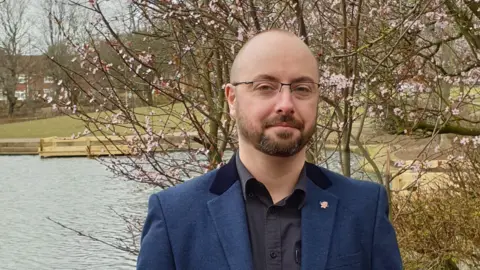 Councillor Tom Feeney is staring at the camera in a black shirt and navy blue suit jacket. There is pond and a pink blossom tree behind him. 