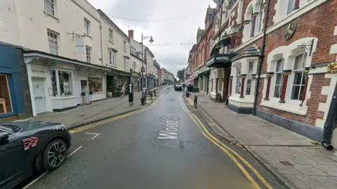A GoogleMaps image of a street with shops either side and a few parked cars. White lettering reads 'Wood Street' on the road.
