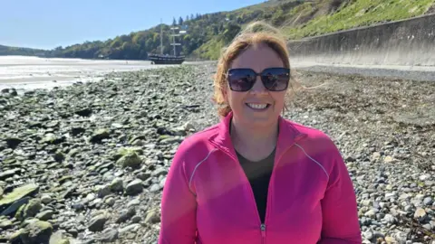 Sinead Fitzpatrick is wearing a bright pink zipped top and has brown hair tied up, and is smiling and wearing sunglasses. She's standing on a rocky part of a beach with a tall ship in the distance on the left, with a tree-lined hilly coastline beyond, on a sunny day with blue skies.