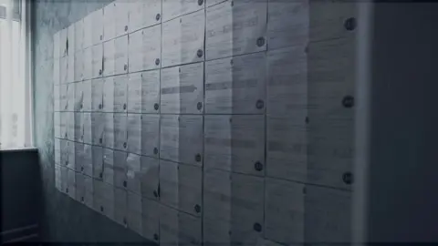 Multiple printed death certificates pinned in neat rows on a wall inside a dimly lit room, with light from a window illuminating the documents and creating vertical shadows.
