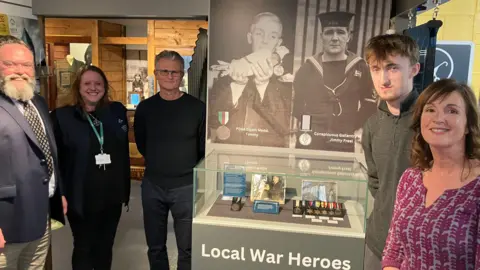 A group of people stand smiling around a museum display case. Medals are in the case, above a sign reading, Local War Heroes. 