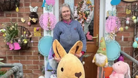 A woman standing in front of a brick house doorway decorated for Easter with colourful eggs, butterflies, hanging baskets and a “Happy Easter” banner, surrounded by plush bunnies and other festive ornaments.