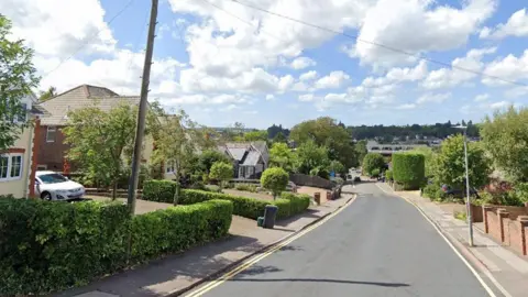 A view of Hillfield Road stretching into the horizon. Houses with large driveways and green trees line each side