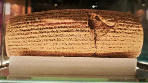A replica of the Cyrus Cylinder in the Parsi museum covered by cuneiform script, displayed in a glass case.