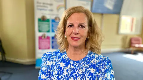 Martin Giles/BBC Caroline Donovan in a bright blue and white dress smiling at the camera. She has long blonde hair and large pearl earrings.