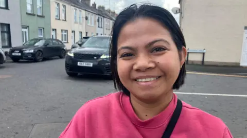 A woman smiles into the camera. She has short black hair and is wearing a pink jumper. She's standing in a street. Cars are parked behind her.
