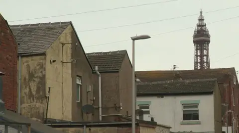 Image of the backs of old terraced houses. The top of Blackpool Tower can be seen poking over the top of one against a grey sky.