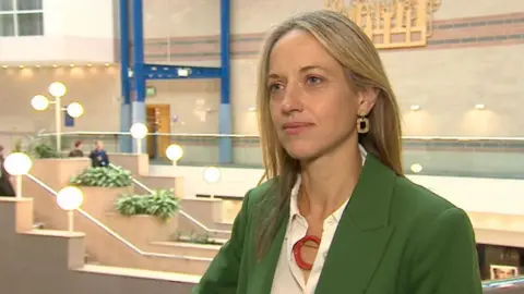 A woman with long blonde hair in a white shirt and green jacket