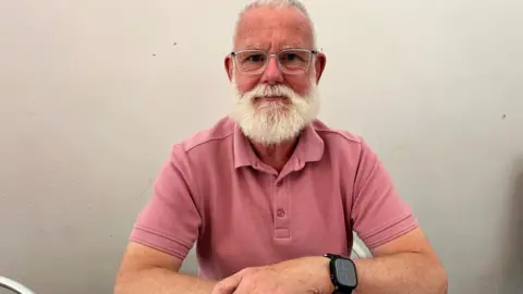 A man with a white beard and glasses sits at a table, wearing a pink polo shirt, with his arms resting in front of him.
