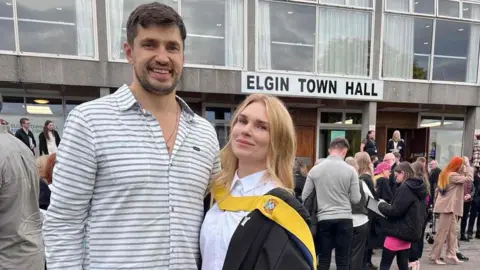 Alex Sivko Alex Sivko, a smiling, bearded young man in a grey and white striped shirt is standing with his wife Inna, who has blonde hair and is wearing a graduation robe with yellow sash and white shirt. They look happy and are standing in front of Elgin Town Hall, which is a grey concrete modernist building.