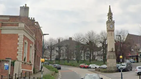 Newnham-on-Severn's high street. A main road passes through with a clock tower on one side and pretty Georgian houses. There are cars parked along the road and around the clock tower Y-junction. Trees missing their leaves can be seen on a village green behind. It is a cloudy day.