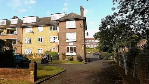 Google street map view of Kingfisher Court, off Queensmere Road, at three-storey brick apartment building with dormer windows and a paved driveway, bordered by grass, shrubs, parked vehicles and mature trees under a blue sky.