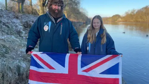 Fenwick Ridley and Frankie Jackson smile as they hold a Union Jack flag. A lake and frost-covered grassland are behind them. They both wear blue with the logo of IISA, the International Ice Swimming Association. 
Frankie has long blonde hair, which is wavy at the ends, blue eyes and dimples. Fenwick has a long brown beard and wears black gloves with open fingers and fur hat covering his ears. 