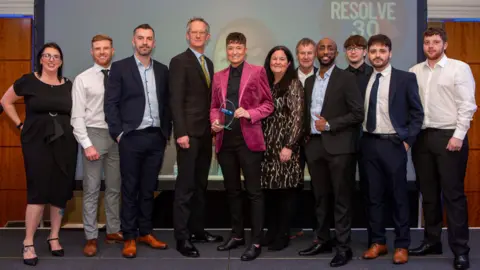 Tiger's Trust Members of Tigers Trust collecting the award. 11 people, smartly dressed, are standing on a stage in front of a big screen. The woman in the centre is holding a glass trophy. 