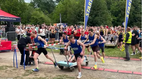 BBC/Oli Constable A team of runners wearing blue tops and black running shorts push a metal trolley between them, with one person sat in the middle. In the background a crowd of people behind a metal barrier can be seen cheering them on. There are two large flags that read: start on either side of the barriers.