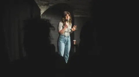 Andrew Max Levy A female comedian stood on stage holding a microphone, silhouetted by members of the crowd.