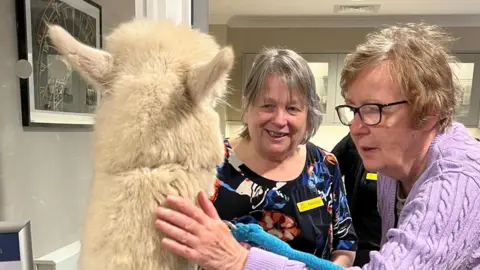 Handout Two women stand with Prancer in the care home and one of them is petting him with her hand on his coat.