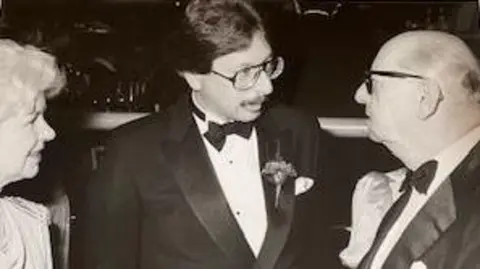 Charles J. Wesoky A black and white photo shows a man dressed in a tuxedo speaking to another smartly dressed man on the right hand side.