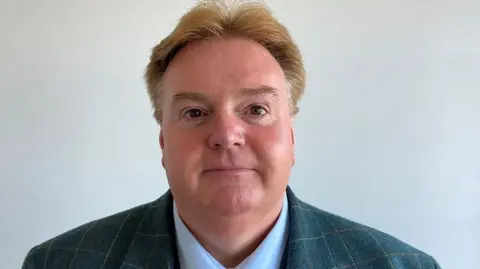 Head and shoulders image of Mark Clifford in front of a plain grey wall. He has sandy hair parted in the middle and is wearing a checked suit and pale blue shirt