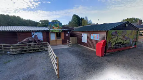 The entrance of a theme park, Hoo Zoo, with colourful signage on a series of single-storey wooden buildings. There is a fence to guide queues and a sign saying "entrance" above a door.