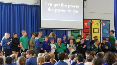 A group of primary school students and athletes stand, and sit underneath a power point which reads "I've got the power, the power in me".