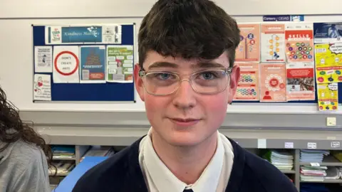 A teenage boy wearing his uniform. He has clear glasses on and has brown hair. School work is hanging up on the walls behind him.