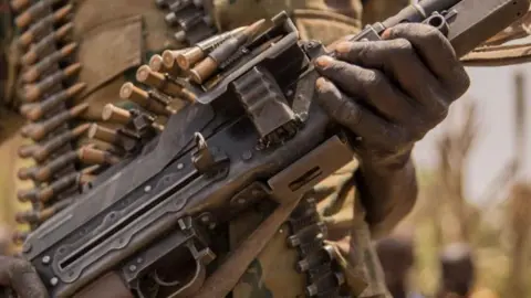 A close up of the body of a gun with a magazine of bullets on top of it. A hand is holding one part of the gun and more bullets can be seen, out of focus, in the background.