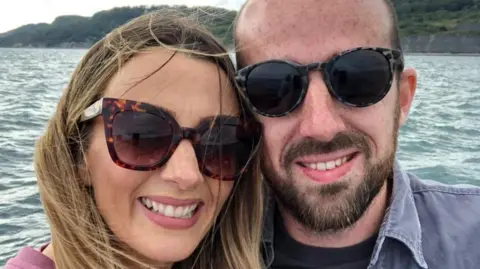 PA Media Oliver and Chloe Long are smiling at the camera. Both are wearing dark sunglasses. She has long, fair hair and he is balding and has a beard and a moustache. There is water behind them and they appear to be on a boat.